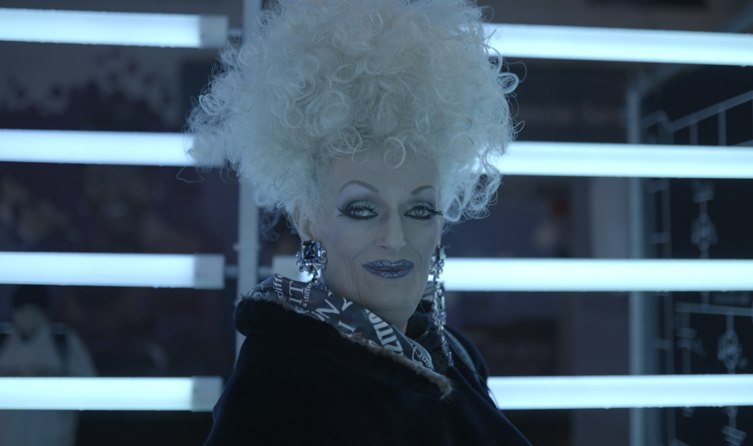 Glamourous woman with glittery makeup, an updo and large dangling earrings wearing a fur lined, collared coat. Drag Icon Oliv Howe.